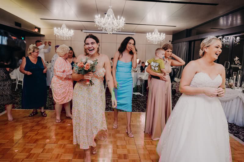 The bride Emily in a white wedding gown laughs on the dance floor at Sandstone Point Hotel — Pumicestone Room while a bridesmaid holding a bouquet walks toward the camera smiling. Other guests, including a woman holding a child and another bridesmaid in a blue dress, are visible in the background.