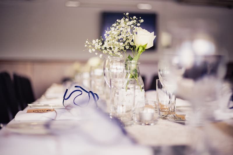 Close-up of a wedding reception table at TOOWONG ROWING CLUB — The Malouf Room, featuring a white rose and baby's breath floral centerpiece in a glass jar, surrounded by glassware, napkins, and place cards.