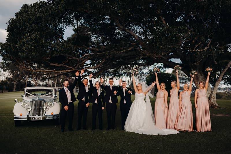 Emily the bride and Dylan the groom pose with their wedding party in front of a vintage white car under a large tree at Sandstone Point Hotel. The bridesmaids wear matching blush pink dresses and hold bouquets raised in the air, while the groomsmen wear black suits with light pink ties.