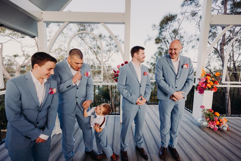 Four groomsmen in light blue suits with pink boutonnieres stand on the pavilion deck at Sandstone Point Hotel, interacting with a young ring bearer dressed in white shirt and suspenders near floral arrangements.