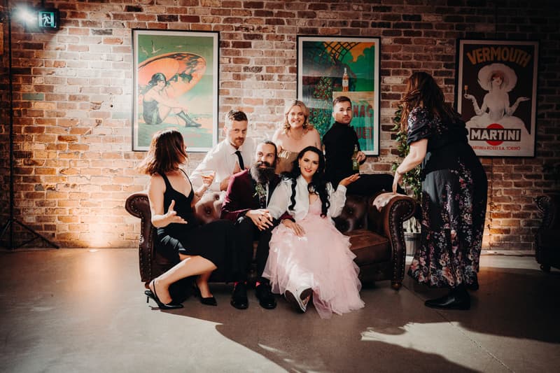 The bride in a pink dress and the groom in a burgundy jacket sit on a brown leather couch at Sandstone Point Hotel — Cellar, surrounded by four guests, two seated and two standing, against a brick wall with vintage posters.