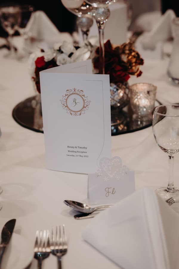 Close-up of a wedding reception table at Royal on the Park featuring a menu card with 'Henny & Timothy Wedding Reception Saturday, 21st May 2022', a decorative place card, silverware, glassware, and floral centerpiece with candles.