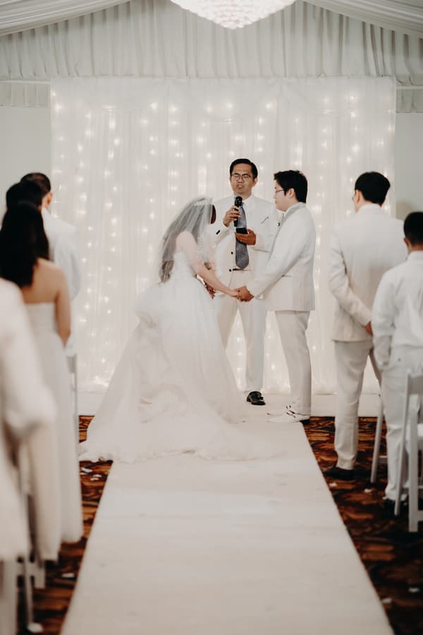 Bride Henny and groom Timothy hold hands facing each other during their wedding ceremony at Royal on the Park, with an officiant speaking into a microphone behind them and guests seated on either side of the aisle.