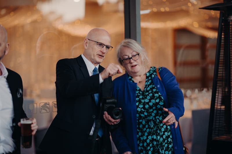 Two older guests, a man in a suit holding a camera and a woman in a blue floral dress and cardigan, are engaged in conversation at The Tides — The Pandanus Room during the wedding reception.