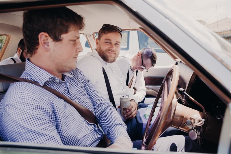 Three men sit inside a car, two wearing white shirts with suspenders and one in a blue checkered shirt. One man holds a can of drink while another appears to be resting with his head down.