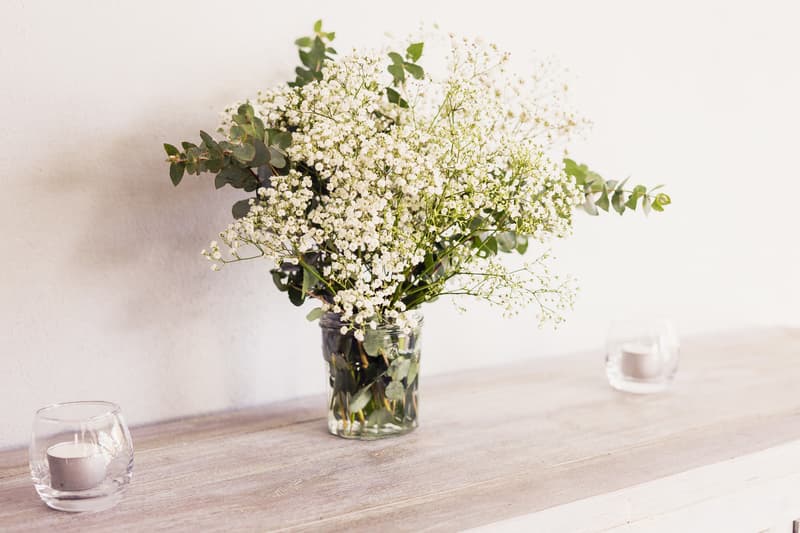 A floral arrangement with white baby's breath and green foliage in a glass jar on a wooden surface, flanked by two small glass candle holders with white candles, at the Toowong Rowing Club — The Malouf Room.