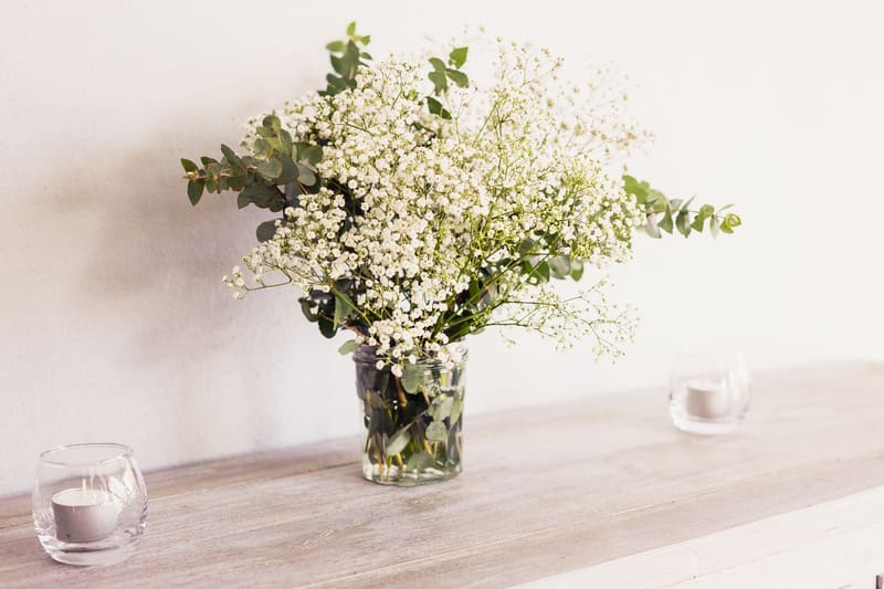 A floral arrangement with white baby's breath and green foliage in a glass jar on a wooden surface, flanked by two small glass candle holders with white candles, at the. What is an engagement party by She Said Yes.