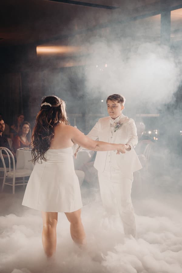 Brooke and Tiffany dance together on the fog-covered floor during their wedding reception at Sandstone Point Hotel — The Pumicestone Room, with seated guests watching in the background.