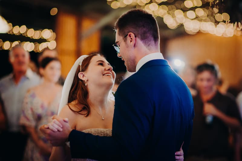 Bride Lilly and groom Connor share a dance at the reception stage of Yabbaloumba Retreat — The Shed with guests blurred in the background and string lights overhead.