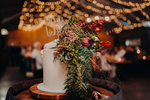 Wedding cake decorated with flowers and a 'Mr & Mrs' topper at the reception stage of Yabbaloumba Retreat — The Shed, with blurred guests and string lights in the background.