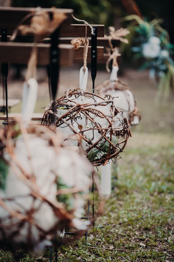 Decorative spherical arrangements made of intertwined branches and white fabric hang from wooden benches at Ocean View Estates ceremony stage.