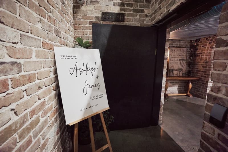 A wedding welcome sign for Ashleigh and James is displayed on an easel against a brick wall near a black door labeled 'CELLAR' at Sandstone Point Hotel — The Cellar.