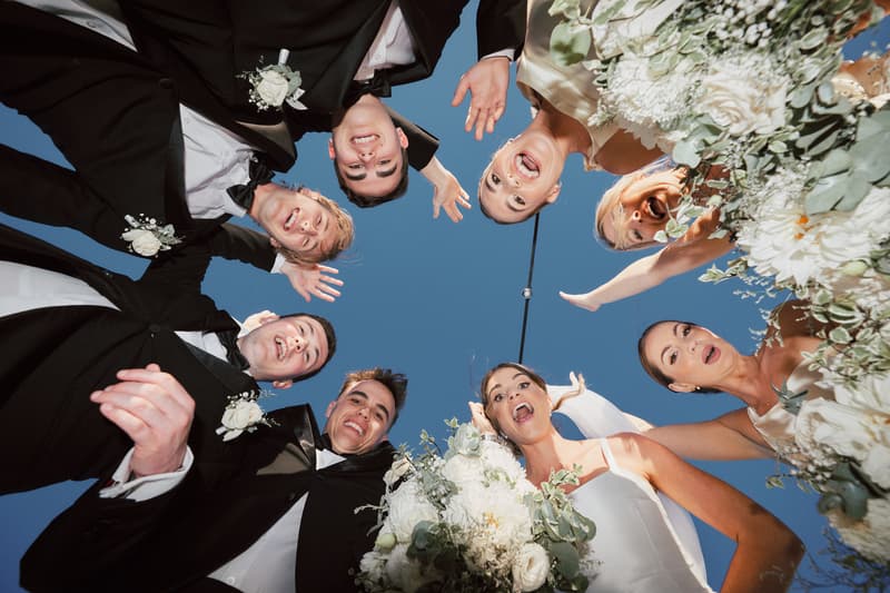 Bride Ashleigh and groom James pose with their bridal party in a circle looking down at the camera at Sandstone Point Hotel, with blue sky background and floral bouquets visible.