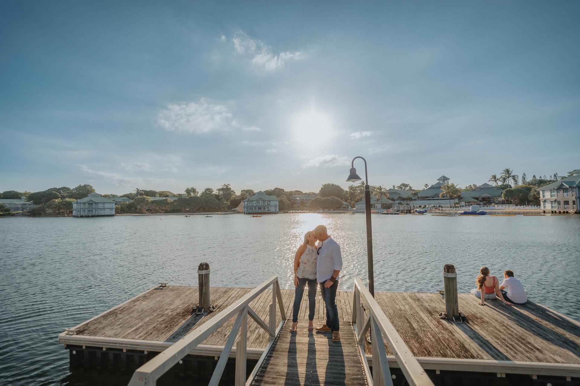 The couple stands on a wooden dock, sharing a tender moment under a bright sky at Novotel Twin Waters.
