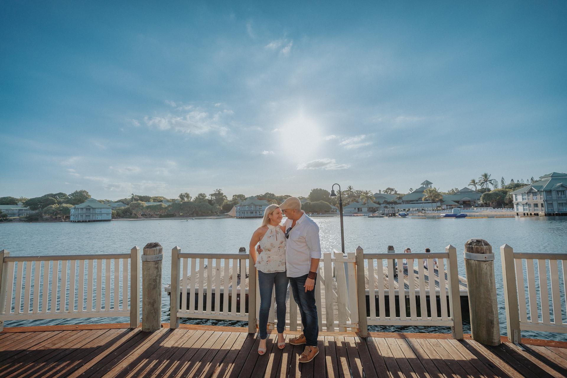 The couple stands close together on a wooden deck, with a scenic water view and clear sky behind them.