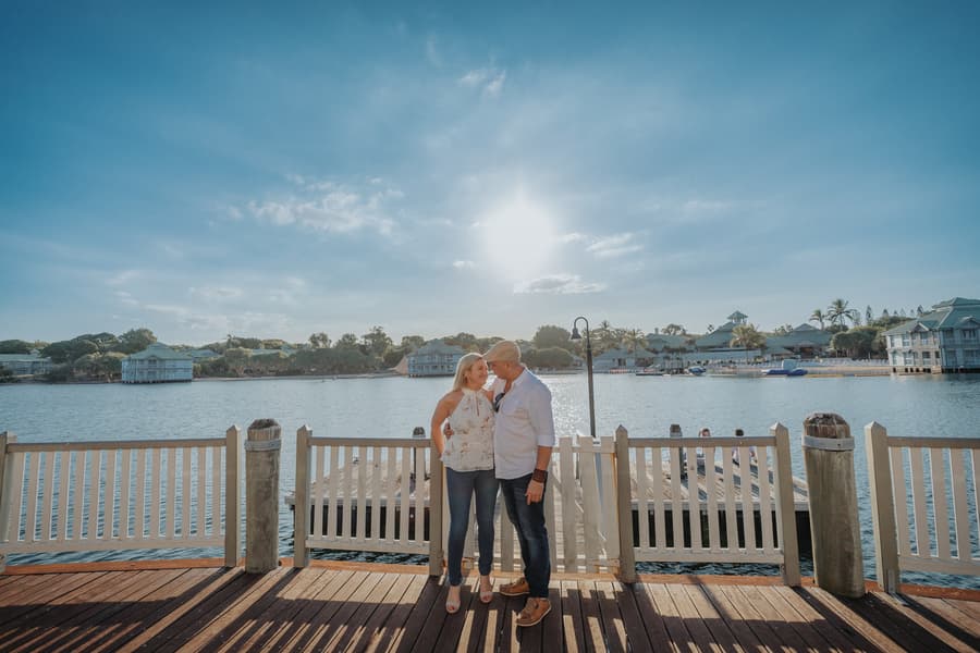 The couple stands close together on a wooden deck, with a scenic water view and clear sky behind them.