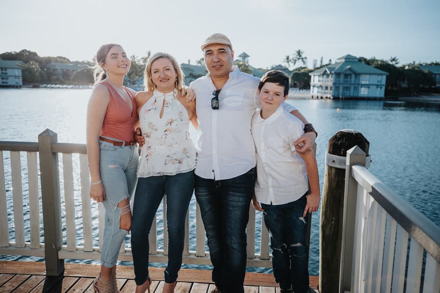 A family of four stands together on a wooden deck, smiling with water and buildings in the background.