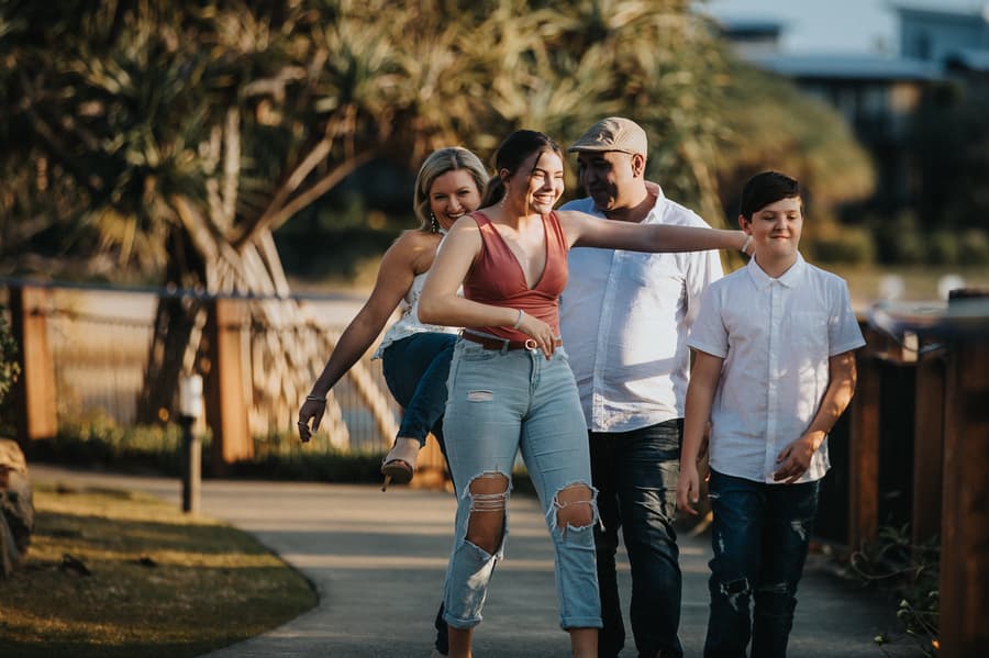 A family of four walks along a path, with one person playfully carrying another, all smiling.
