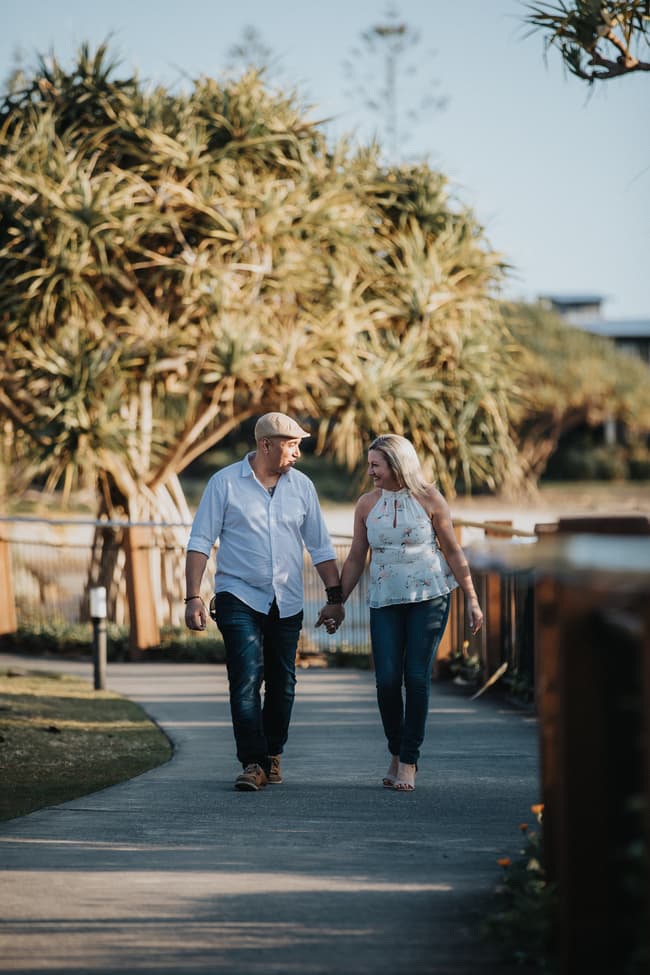 The couple walks hand in hand along a path, surrounded by trees and greenery at Novotel Twin Waters.