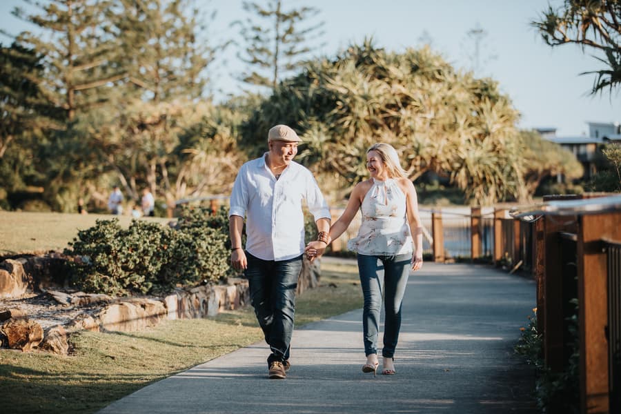 The couple walks together on a path, holding hands and smiling, with trees lining the way.