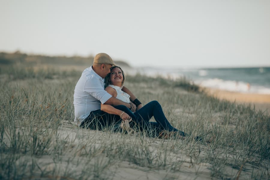 The couple sitting on the beach, the man whispering to the woman, both smiling warmly.
