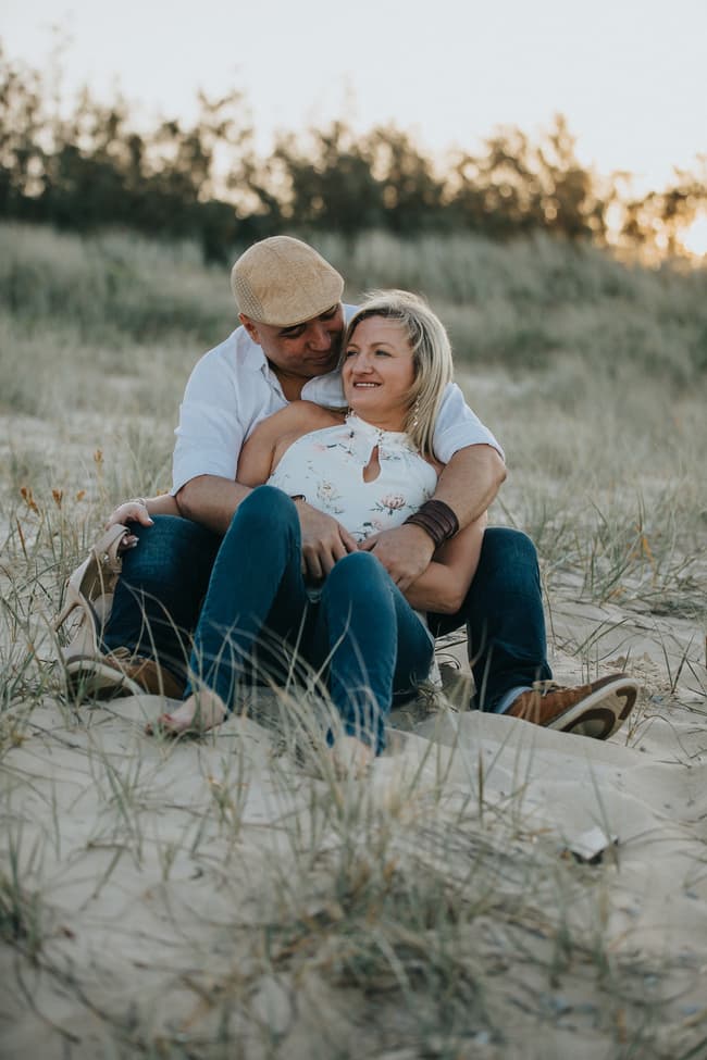 The couple sitting on the sand, the woman holding her shoes, both looking content and relaxed.