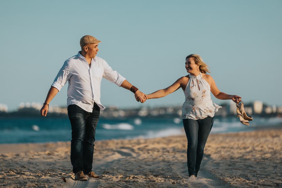 The couple walking hand in hand along the beach, with the woman holding her shoes, under a clear blue sky.