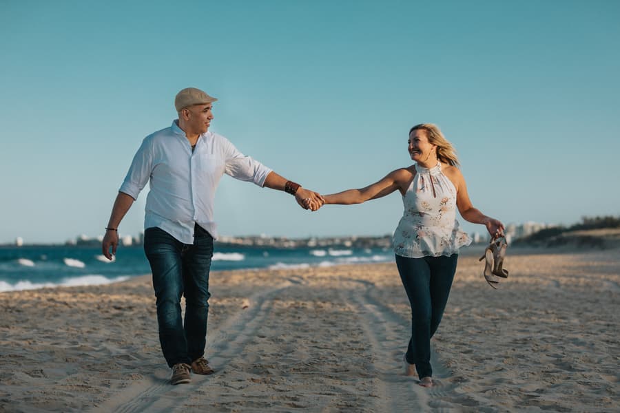 The couple walking along the beach, holding hands and smiling, with waves in the background.
