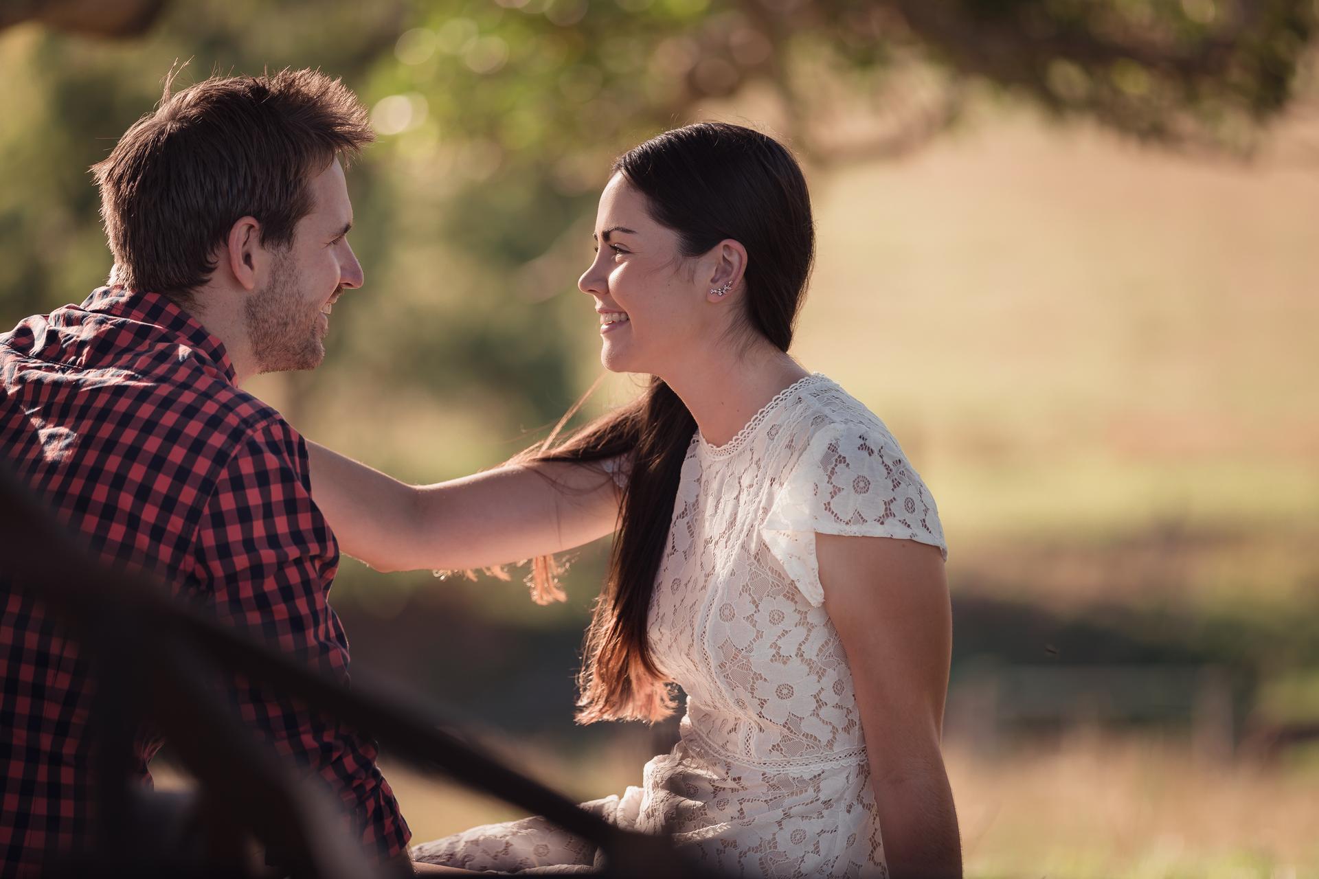 Close-up of the couple smiling at each other under a tree, with soft sunlight filtering through the leaves.