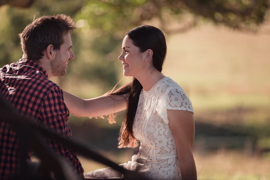 Close-up of the couple smiling at each other under a tree, with soft sunlight filtering through the leaves.