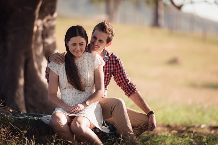 The couple sits together on the grass, with one leaning against the other, both smiling in a sunlit field.