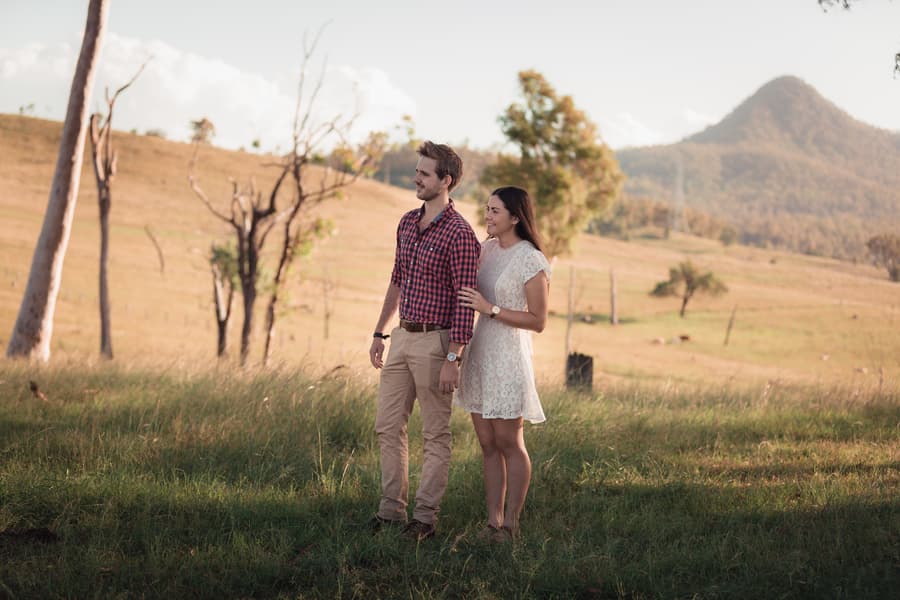 The couple stands in a grassy field, looking into the distance with trees and hills in the background.