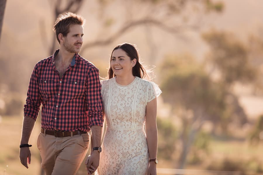 The couple walks hand in hand through a sunlit field, smiling and enjoying the moment.