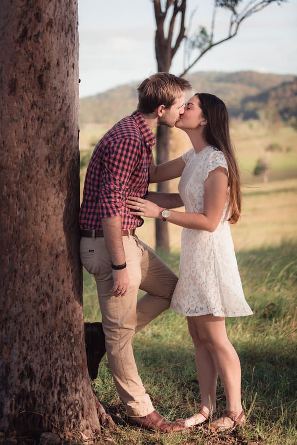 The couple shares a kiss while leaning against a tree, surrounded by a lush green landscape.