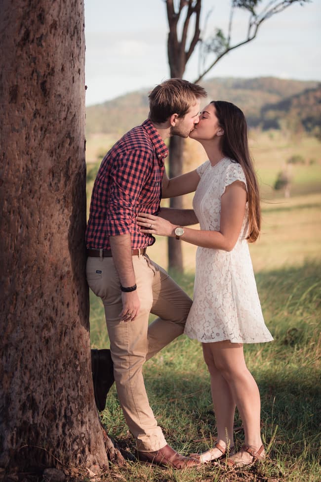 The couple shares a kiss while leaning against a tree, surrounded by a lush green landscape.