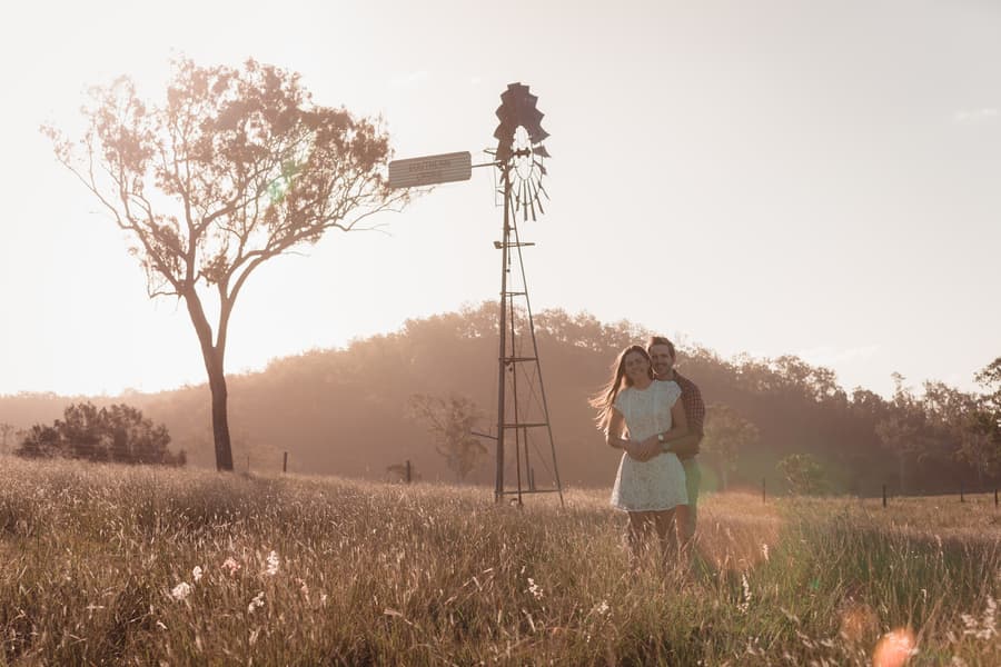 The couple embraces in a field with a windmill in the background, bathed in warm sunlight.