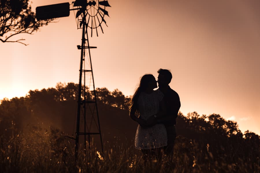 Silhouette of the couple embracing in a field at sunset, with a windmill in the background.