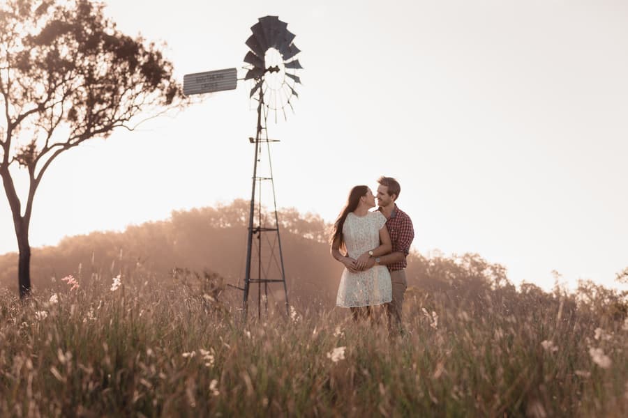The couple stands together in a field with a windmill, surrounded by soft morning light.