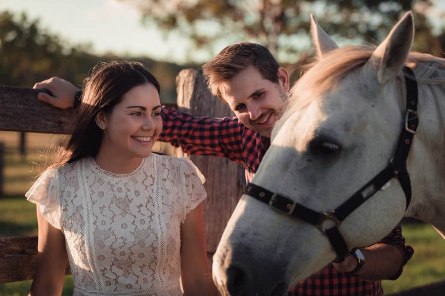 The couple smiling beside a white horse, leaning on a wooden fence at a farm near Ipswich.