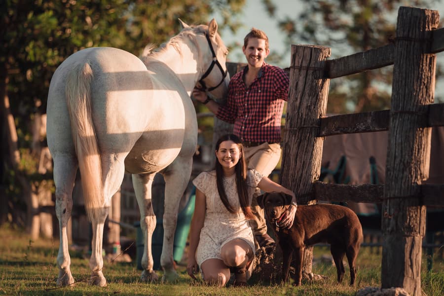 The couple with a white horse and a brown dog, smiling near a wooden fence at a farm.