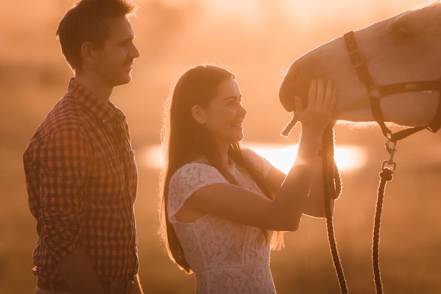 The couple standing close, the woman touching the horse's nose in a golden sunset glow.