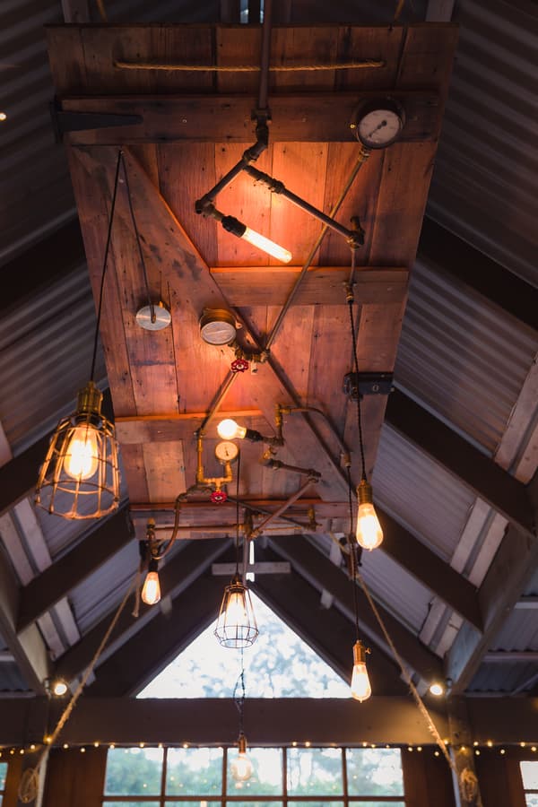 Ceiling view at Kwila Lodge showing rustic wooden panel with industrial-style hanging light bulbs and metal pipes, inside a building with a peaked roof and large windows.