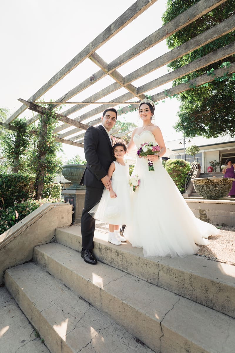 The groom, bride, and a young girl in a white dress pose together on steps under a wooden pergola. Winter wedding light photography Brisbane by She Said Yes.