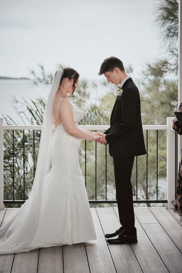 Terion the bride and Tyler the groom hold hands facing each other during their wedding ceremony at Sandstone Point Hotel — The Pavilion, with a waterfront and greenery visible in the background.