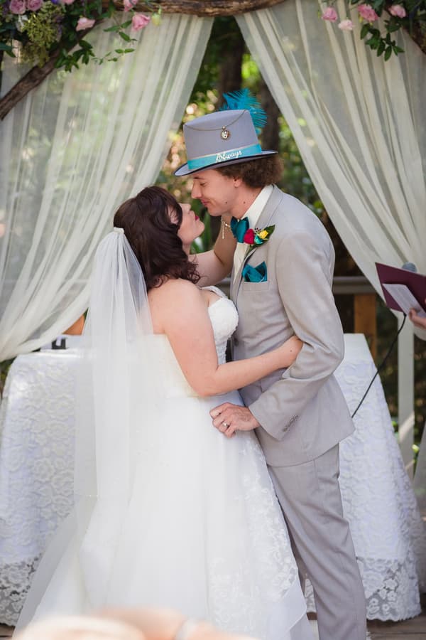Bride Holly and groom Paul embrace and lean in for a kiss at the ceremony stage at Kwila Lodge, standing under a floral arch with white draped curtains.