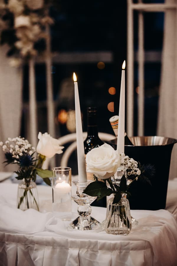 Table centerpiece at the Sandstone Point Hotel Pumicestone Room reception featuring two lit white taper candles in glass holders, white roses in small glass vases, a lit votive candle in a glass holder, a wine bottle, and a black ice bucket.