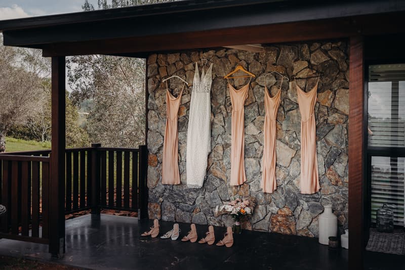 The bride's white wedding dress and four bridesmaids' peach dresses hang on a stone wall at Preston Peak Winery Functions Centre, with matching shoes and a bouquet arranged below.