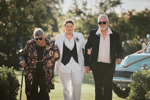 Bride Selina walks arm-in-arm with an older woman and an older man, likely her parents, outdoors, with a vintage car visible in the background. Wedding photographer and videographer by She Said Yes.