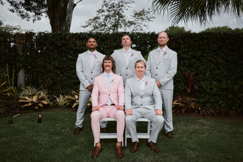 Jake in a pink suit and four groomsmen in light grey suits pose for a group portrait outdoors at Eatons Hill Hotel.
