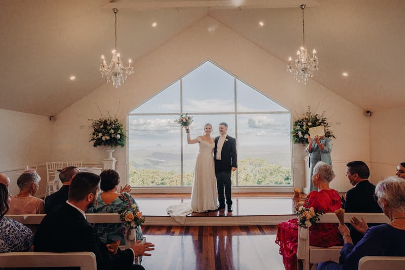Courtney and Liam stand on the ceremony stage at Tiffany's Maleny — Chapel, with Courtney holding a bouquet aloft. Guests are seated on white benches facing the couple, and a person stands to the right holding a certificate. The large window behind the couple shows a scenic view of hills and sky.